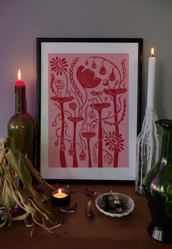 Framed surreal floral art print in red and pink tones displayed on a cozy altar-like setup with lit candles, dried herbs, a green bottle, and a white wax-dripped candle. A warm, mystical ambiance enhances the print’s botanical fantasy style.