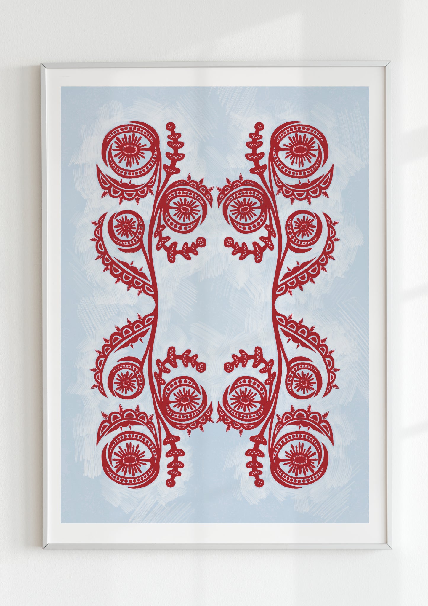 Framed folk-inspired red and blue wall art print with symmetrical floral and spiral motifs, displayed in a white frame on a light background, Scandinavian or Slavic aesthetic.