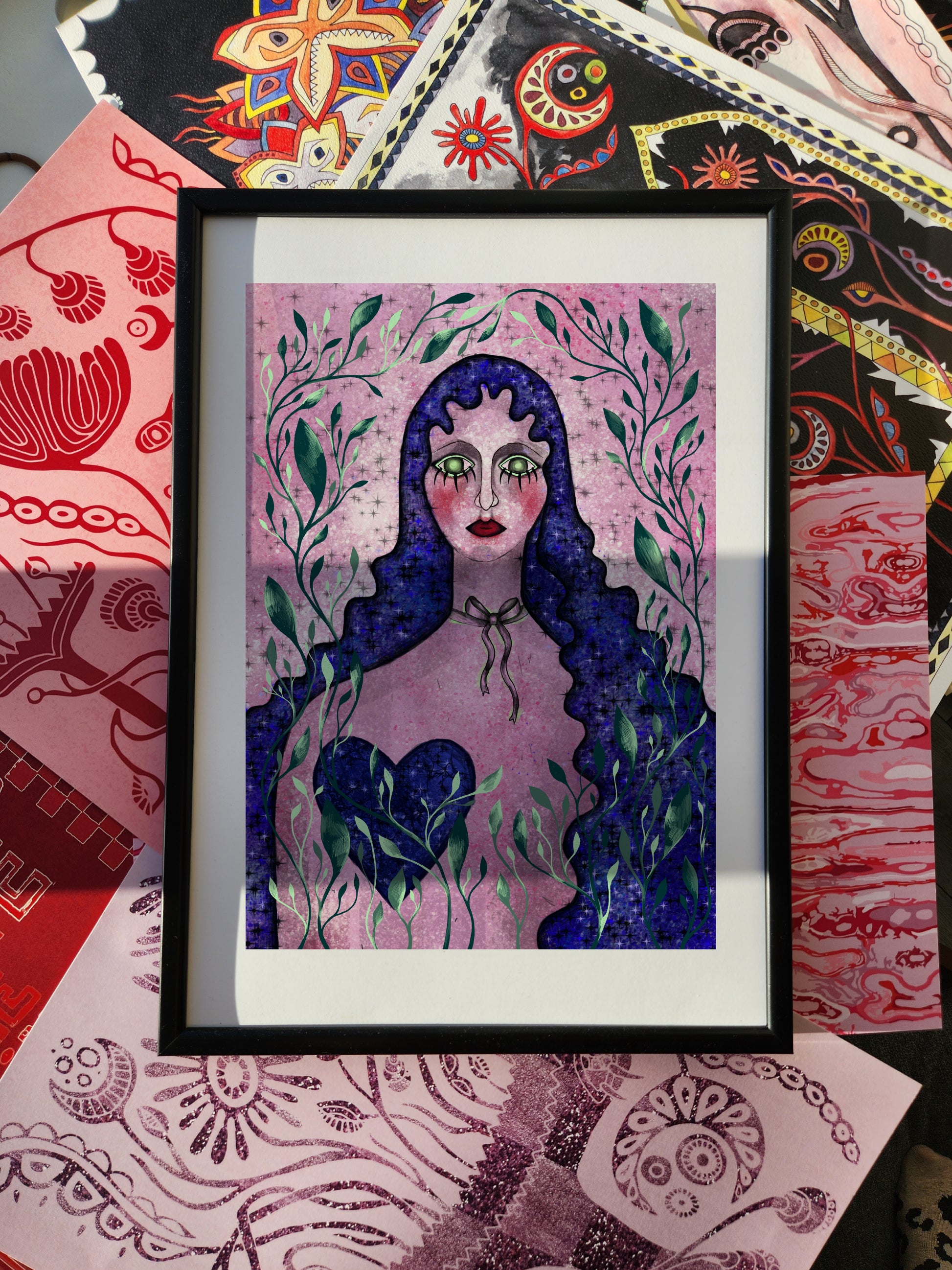 Pink wall art portrait poster featuring a surreal female figure with dark blue hair, symbolic heart motif, and floral decorative elements, framed against an eclectic patterned background.